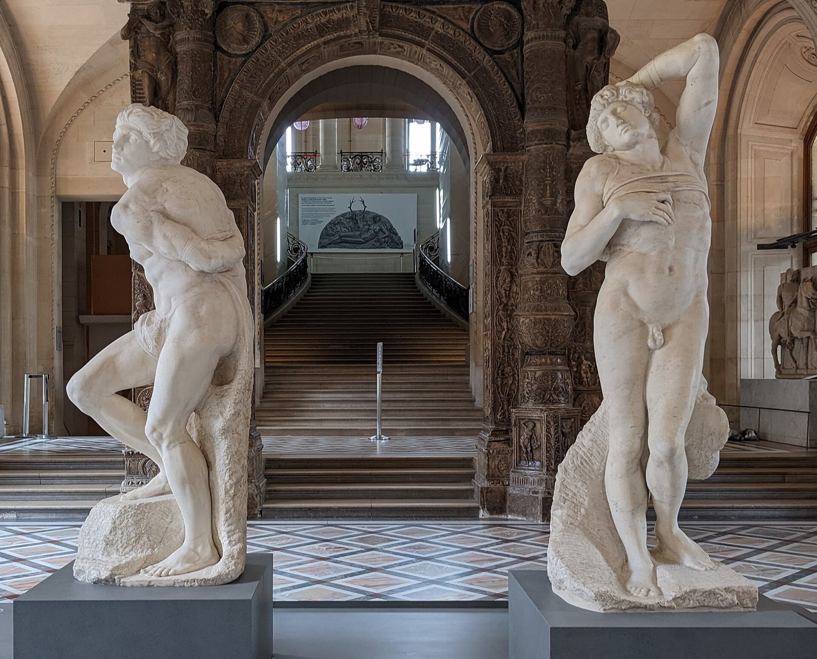 Two marble Slaves by Michelangelo in a gallery of the Louvre: one twists against his bonds, the other leans back with an arm raised behind his head and his eyes closed.
