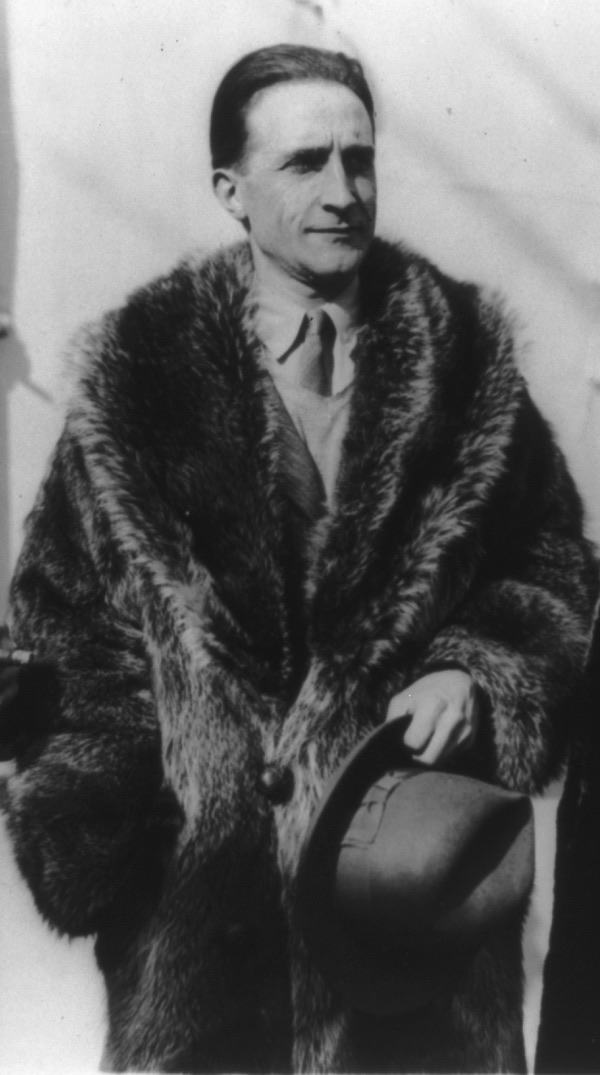 A black-and-white photograph of Marcel Duchamp in a heavy raccoon fur coat, holding his hat and looking off to one side.